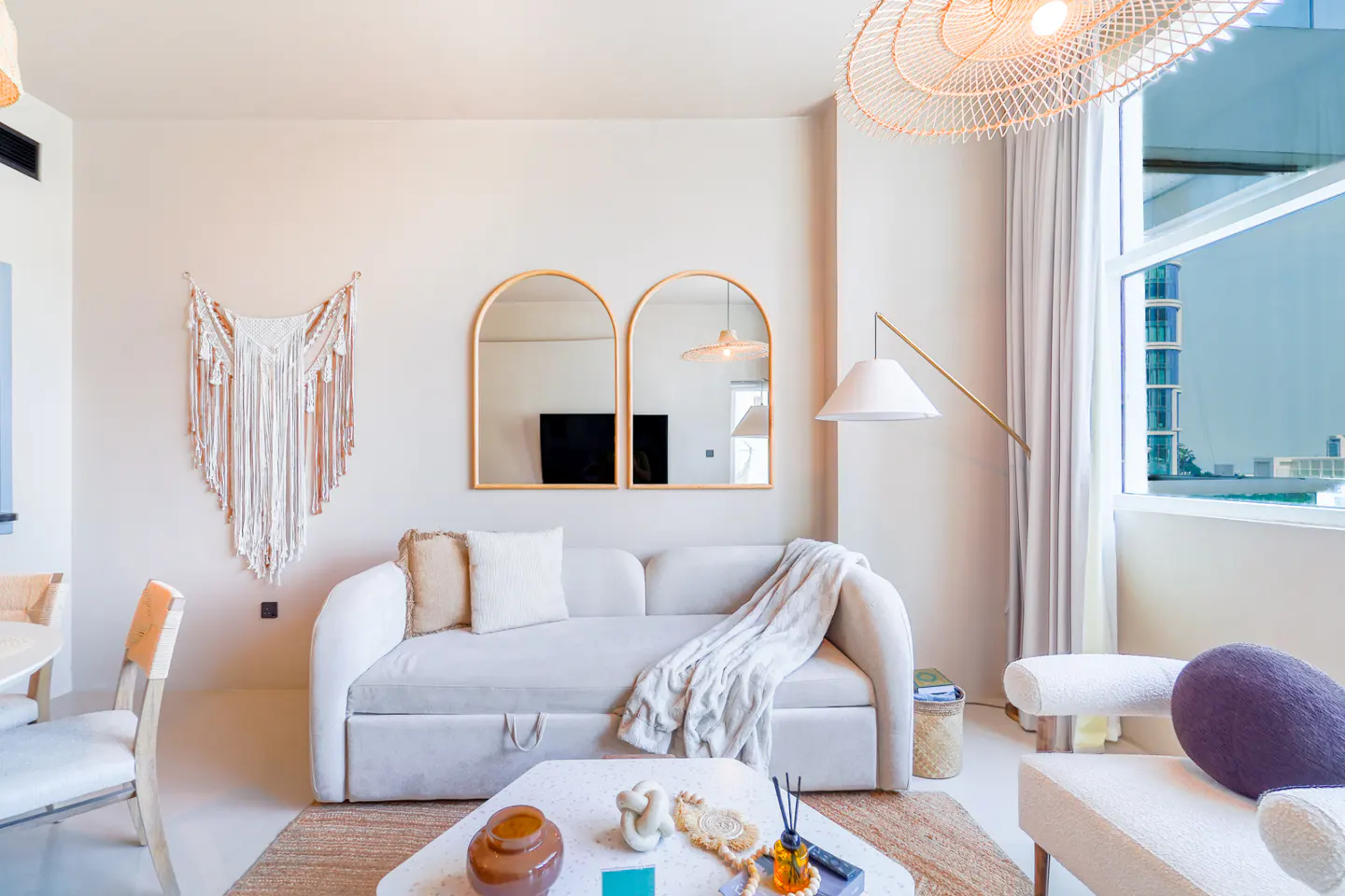 Living room with beige walls, white sofa, and arched mirrors. A macrame wall hanging and a rattan chandelier add texture. Large window with city view.