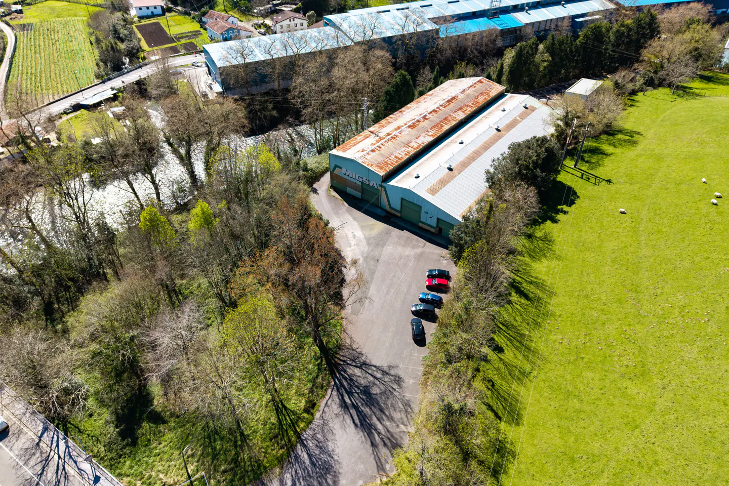Aerial view of a metal warehouse with a rusty roof, surrounded by green fields, trees, and a river. Several cars are parked in front.