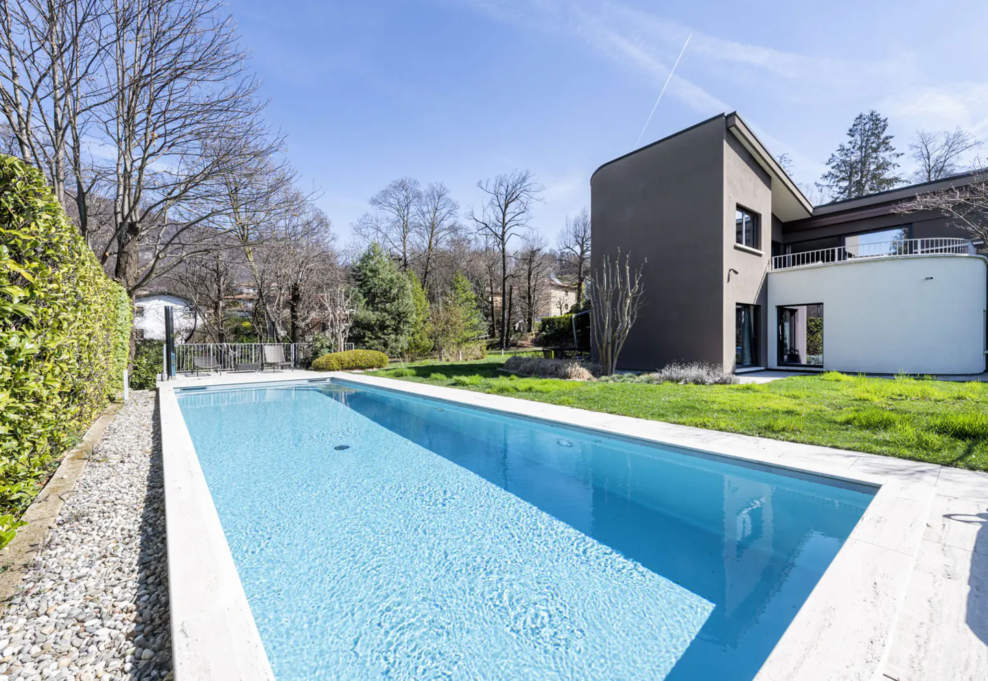 A long, rectangular swimming pool with clear blue water sits beside a modern, two-story house with a gray exterior and white accents.