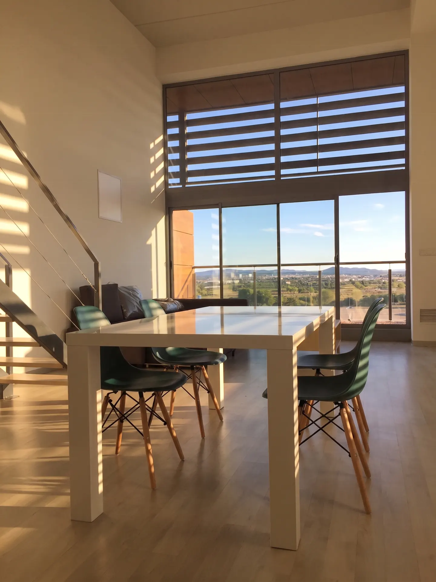 Modern dining area with a white table, green chairs, and large windows overlooking a landscape at sunset.