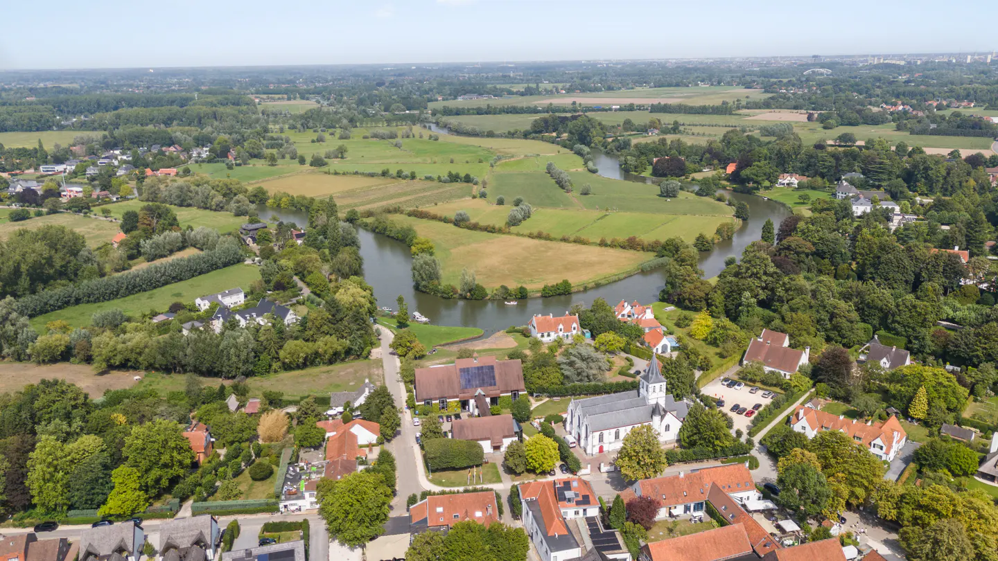 Aerial view of a village with red-roofed houses, a white church, and a winding river through green fields.