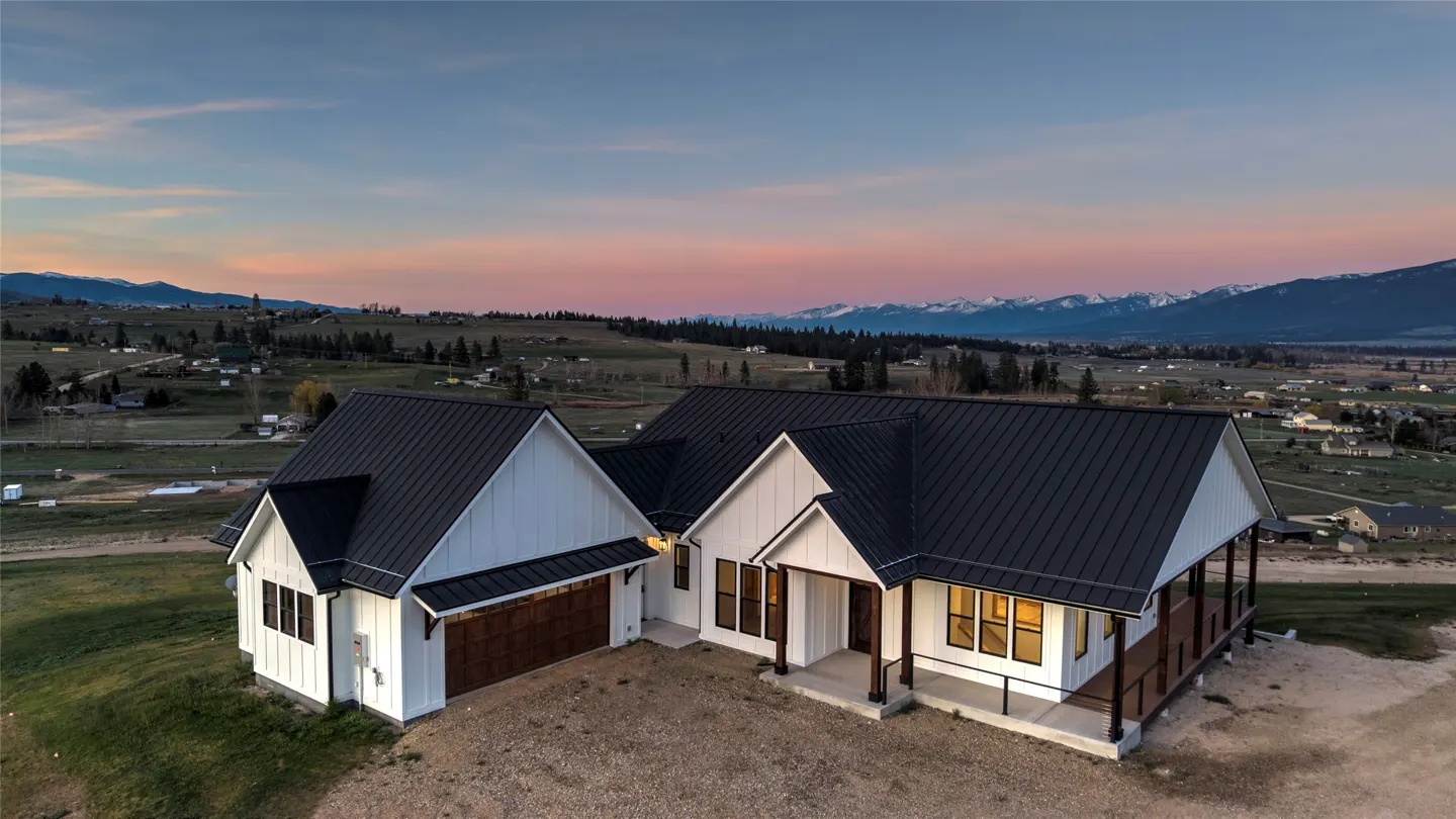 Aerial view of a modern white farmhouse with a black roof, set against a mountain backdrop at sunset.