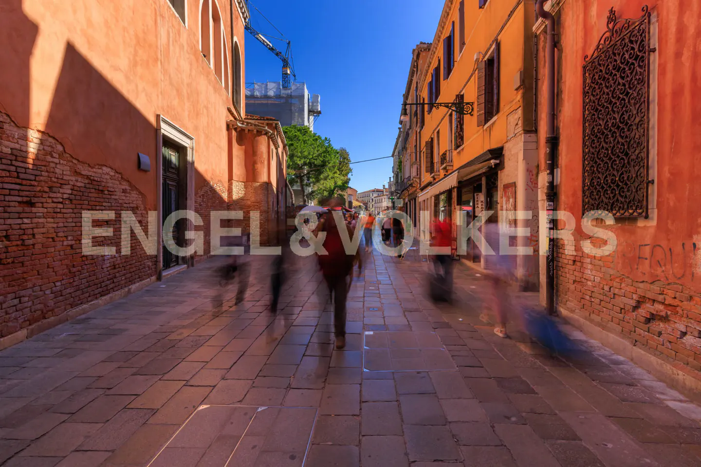 A narrow street in Venice, Italy, with orange buildings on either side and blurred figures walking along the stone-paved path.