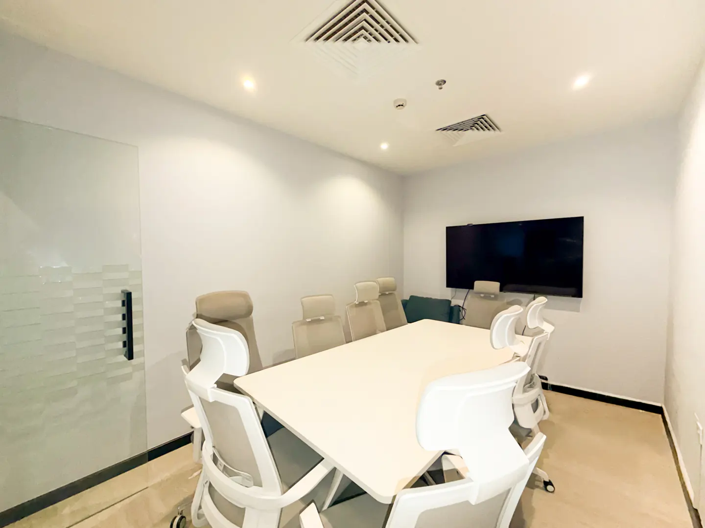 Modern meeting room with a white table, six ergonomic white chairs, and a large wall-mounted television screen.