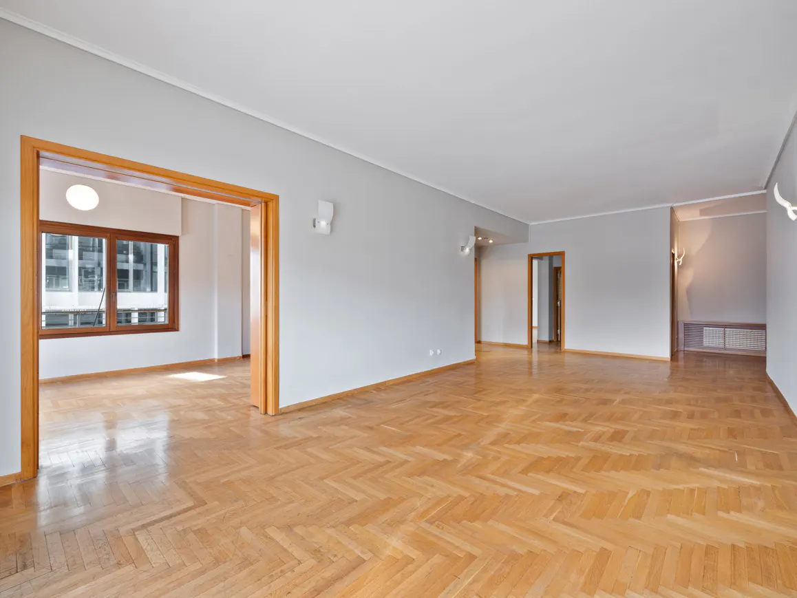 Empty room with herringbone wood floors, gray walls, and white ceiling. Doorways lead to other rooms. A window is visible in the distance.