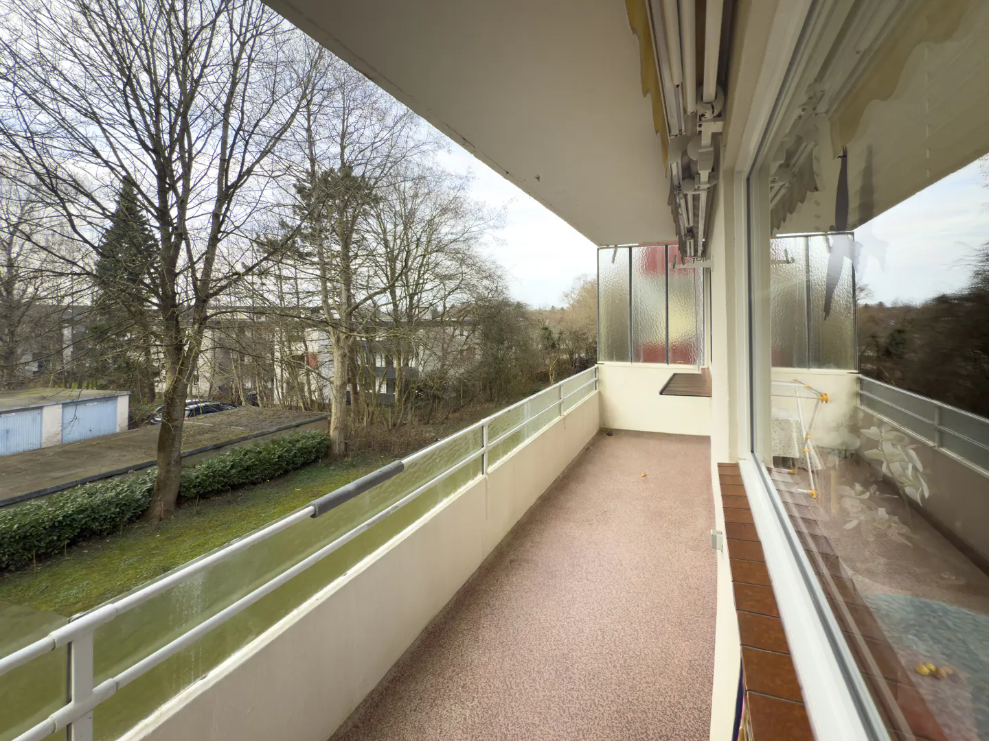 A balcony with a white railing overlooks trees and buildings. The balcony has a speckled brown floor and a white ceiling.