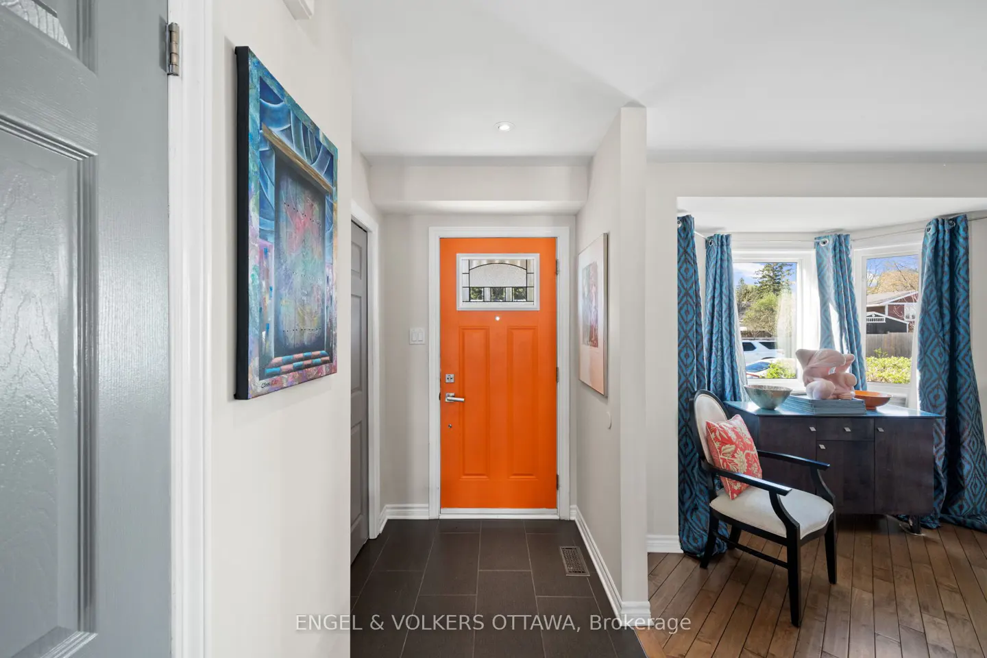 A bright orange front door with a glass panel is centered in a home's entryway. A gray door is to the left, and a living room is to the right.