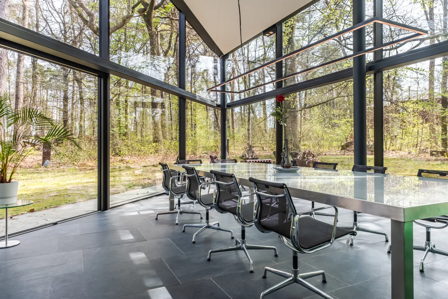 Modern dining room with floor-to-ceiling windows overlooking a forest. A long metal table is surrounded by black chairs.