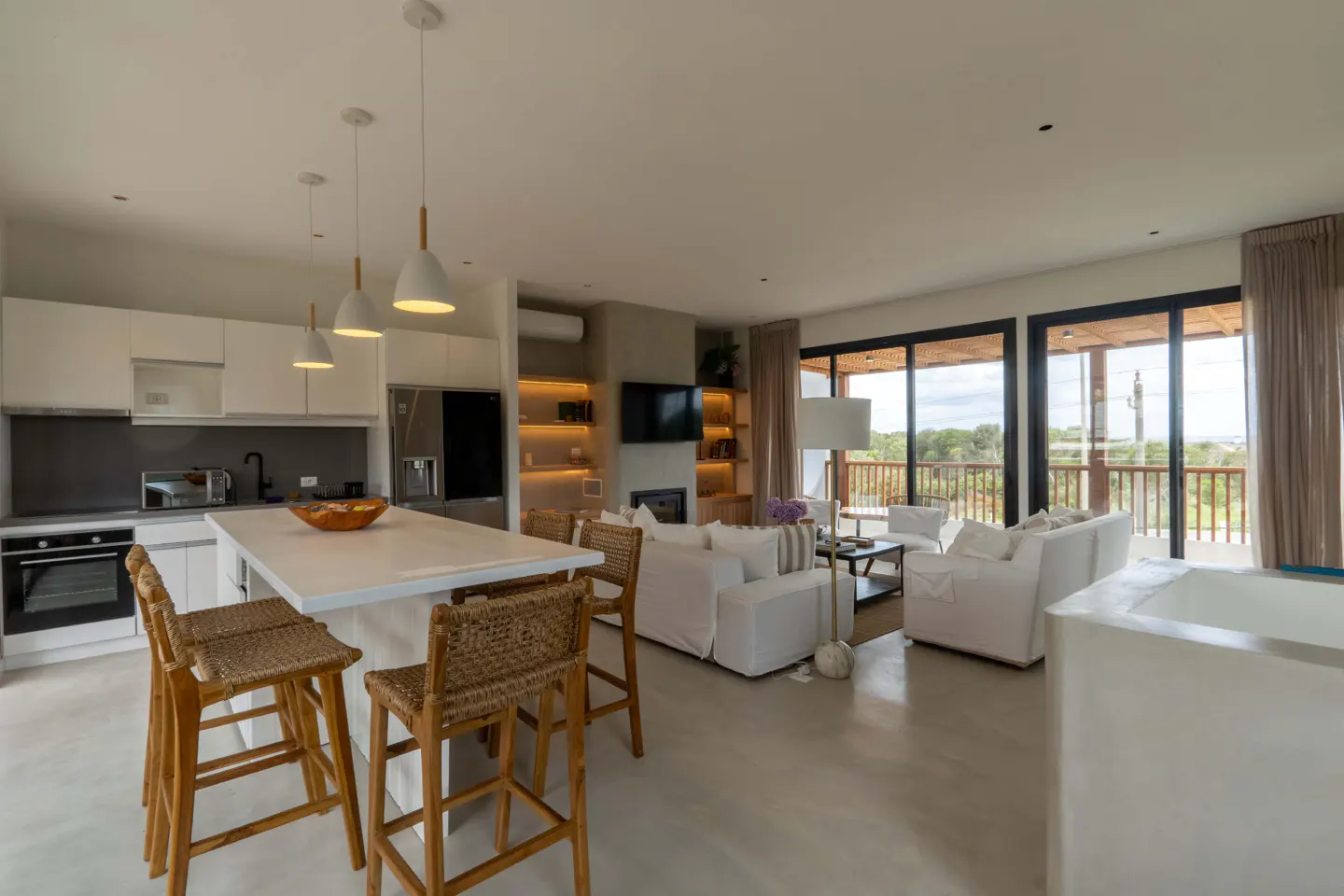 Open-concept living space with white kitchen, island with wicker stools, and white sofas facing a TV and fireplace. Balcony view.