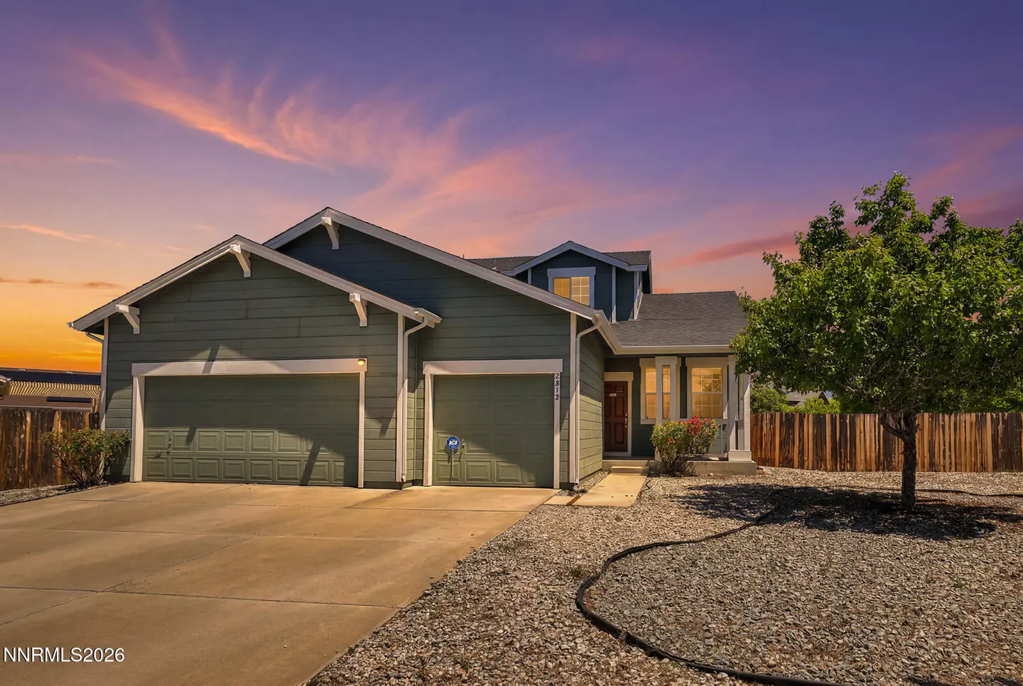 Green two-story house with a two-car garage and gravel front yard under a vibrant sunset sky.