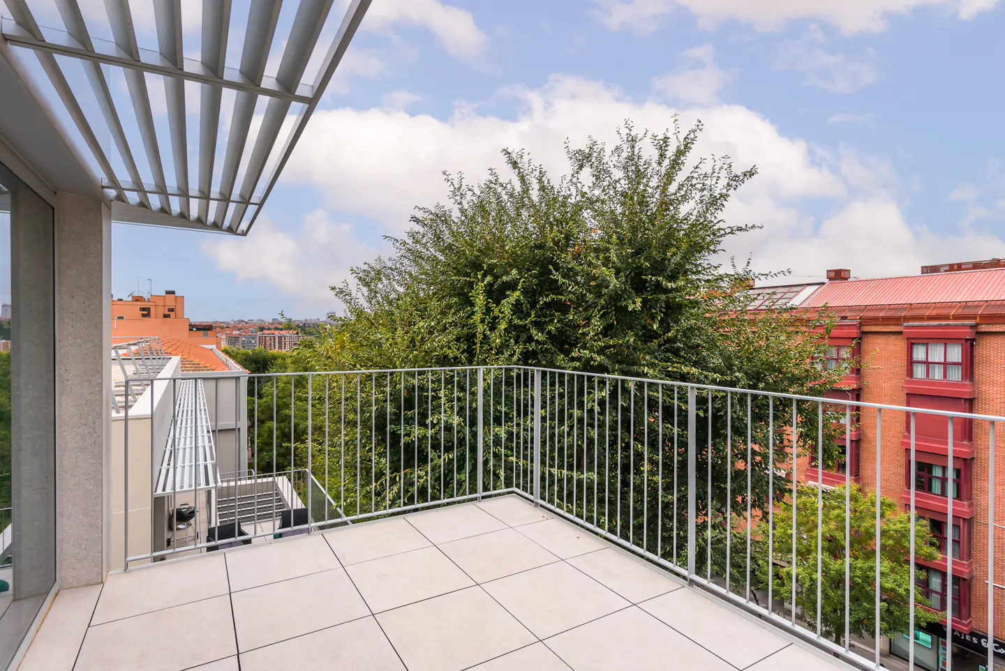 A bright balcony with white tile flooring and a metal railing overlooks trees and red brick buildings under a cloudy sky.