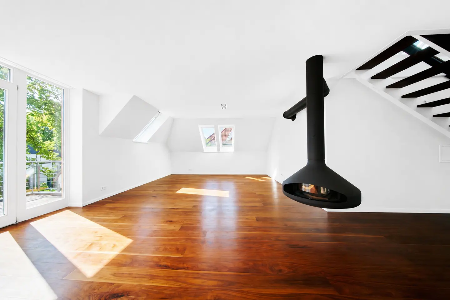 Bright, empty room with hardwood floors, white walls, and a modern black hanging fireplace. Windows and a staircase are visible.