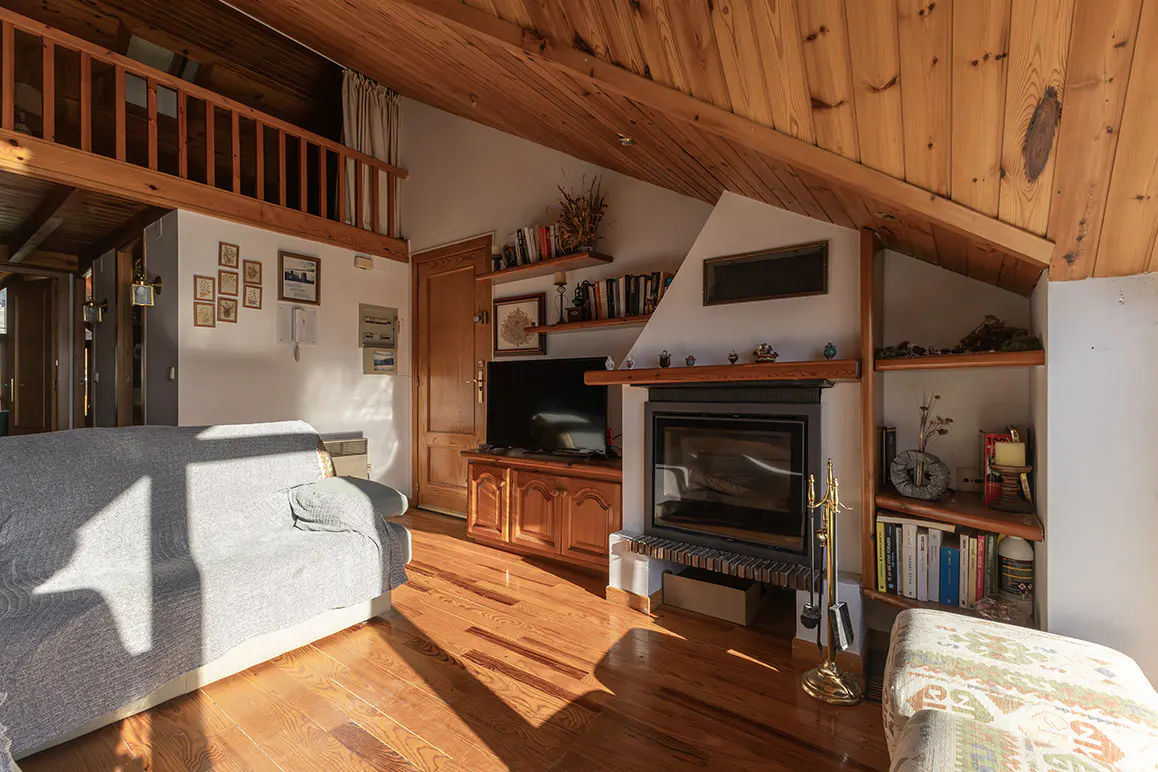 Living room with wood floors, a gray sofa, and a fireplace. The ceiling is wood and there is a loft area.