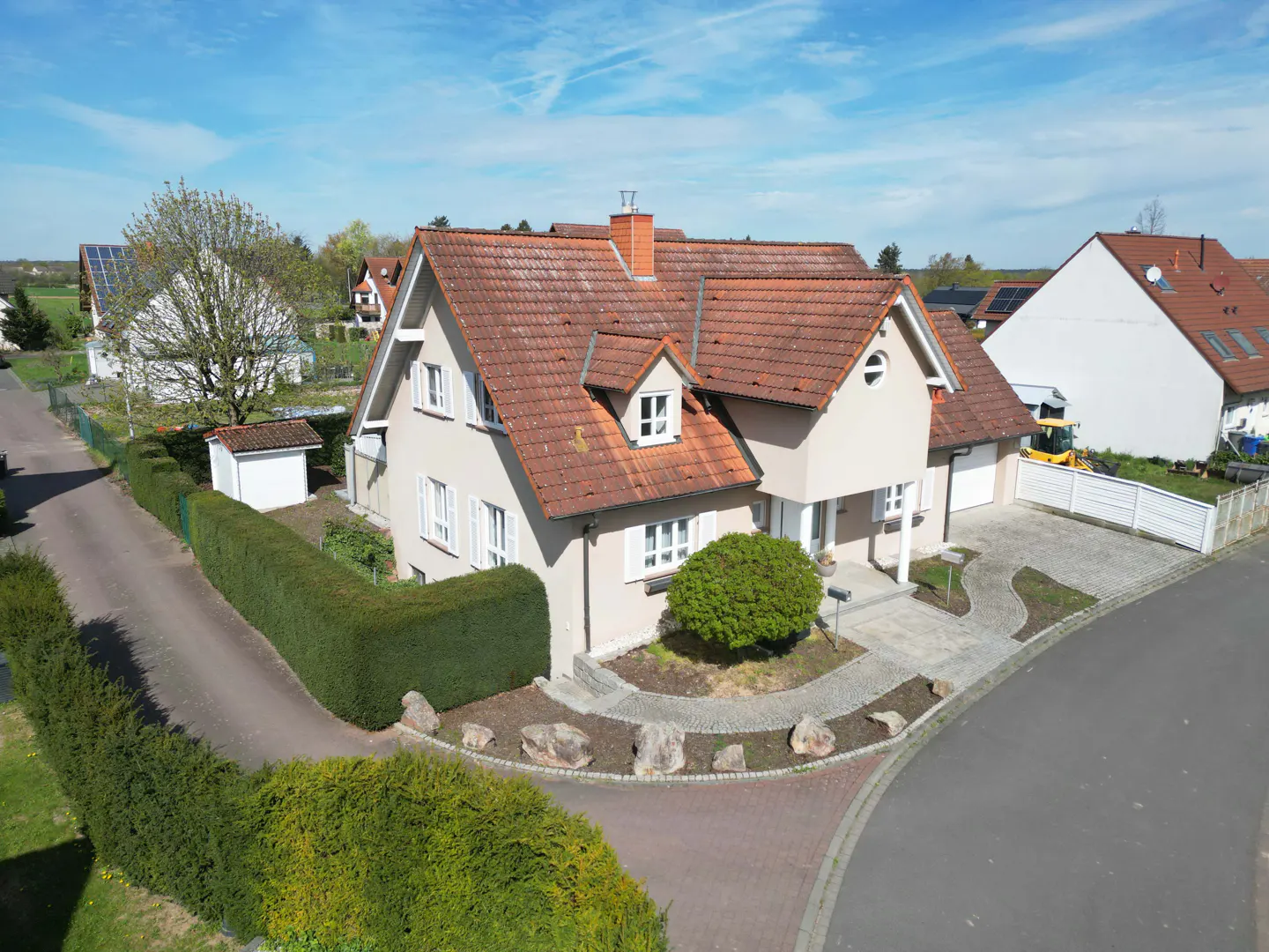 Beige two-story house with a red tile roof, white shutters, and a green hedge in the front yard on a sunny day.