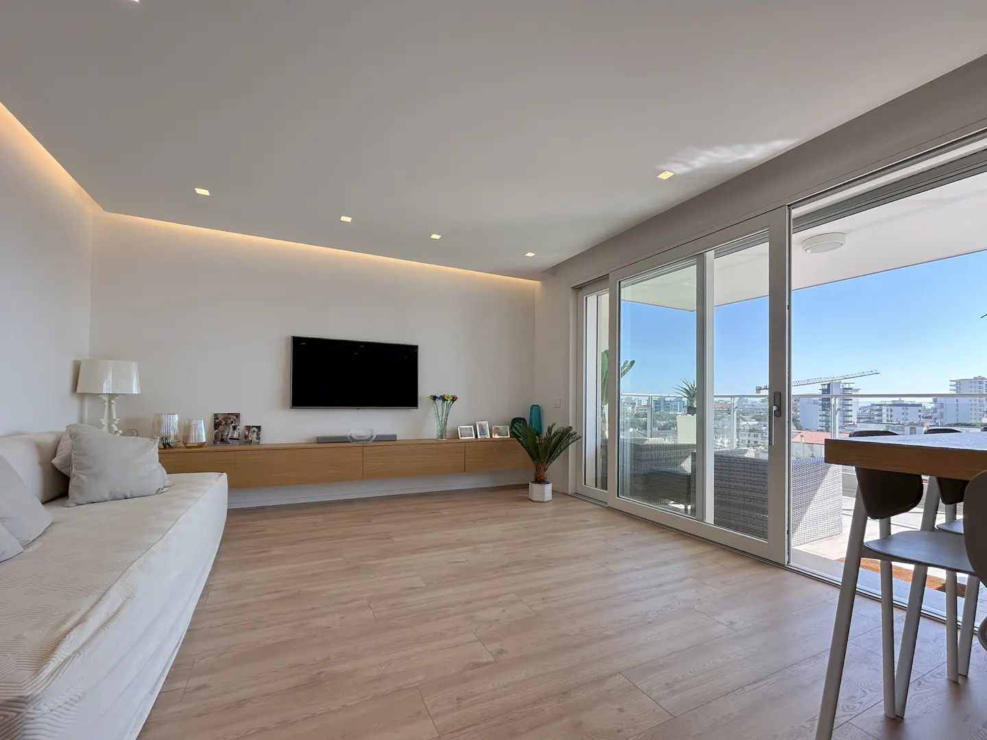 Bright living room with wood floors, white walls, and recessed lighting. A large sliding glass door leads to a balcony with city views. A TV hangs above a light wood console.