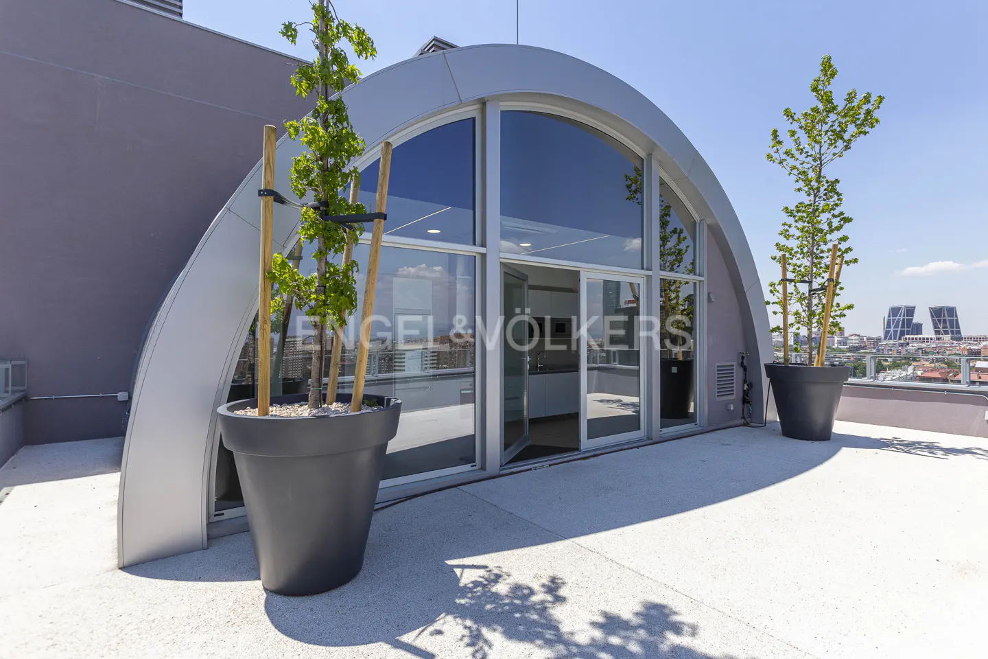 Modern rooftop terrace with arched glass doors, potted trees, and city views on a sunny day.