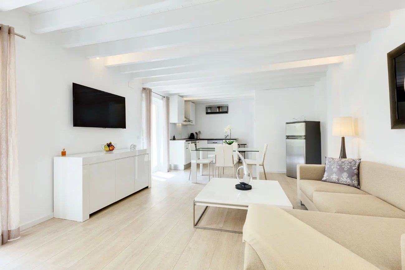 Bright, open-concept living space with white walls, wooden floors, and exposed beams. A beige sofa faces a white coffee table and TV. Kitchen in background.