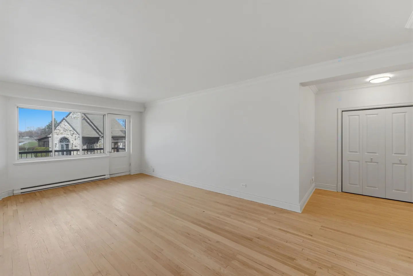 Bright, empty room with light wood floors, white walls, and a large window showing a stone building outside. A white closet door is on the right.