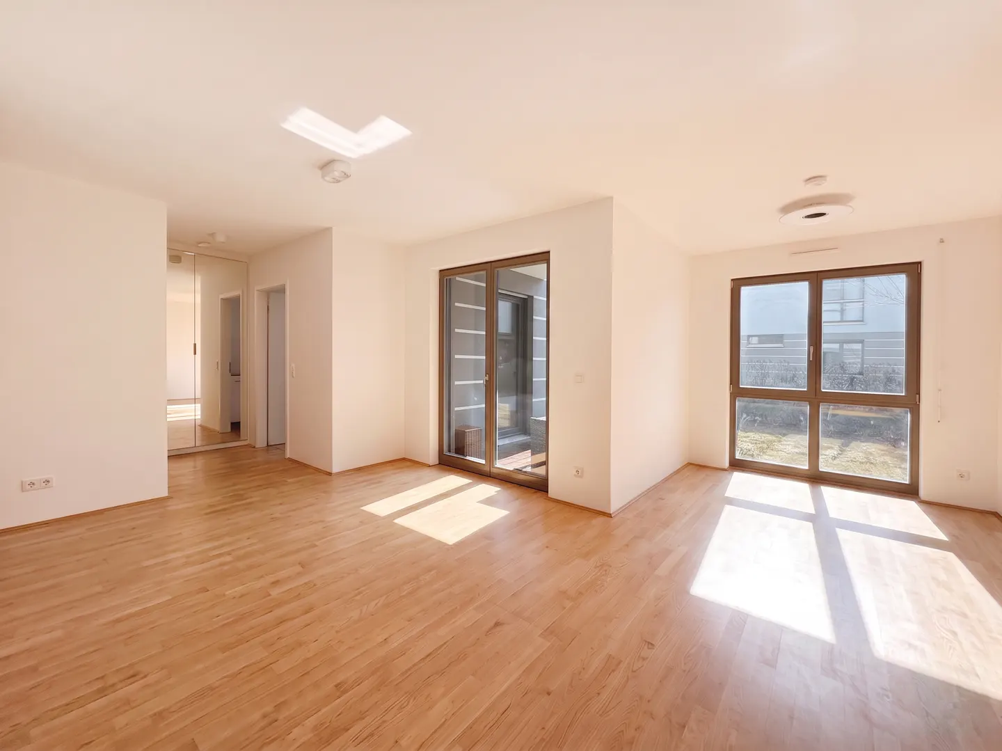 Bright, empty room with light wood floors, white walls, and two large windows. Sunlight streams in, highlighting the spacious interior.