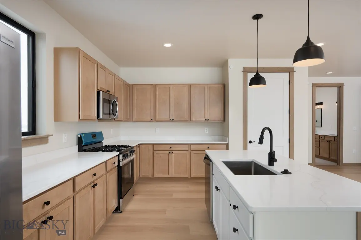 Bright kitchen with light wood cabinets, white countertops, and stainless steel appliances. Black pendant lights hang above a white island.