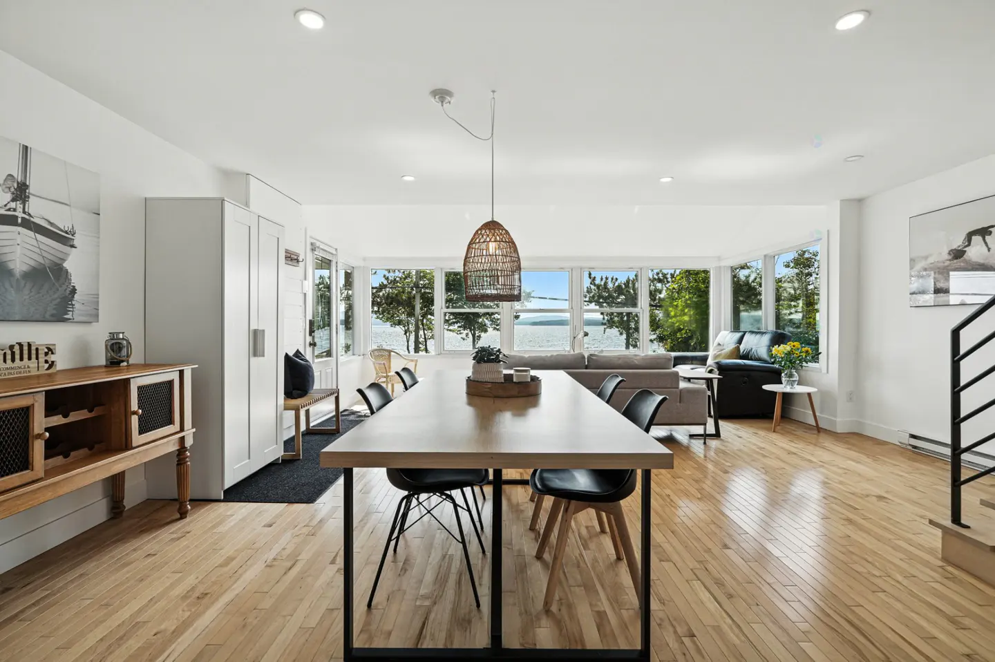Bright, open-concept living space with a long wooden table, black chairs, and a view of the water through large windows.