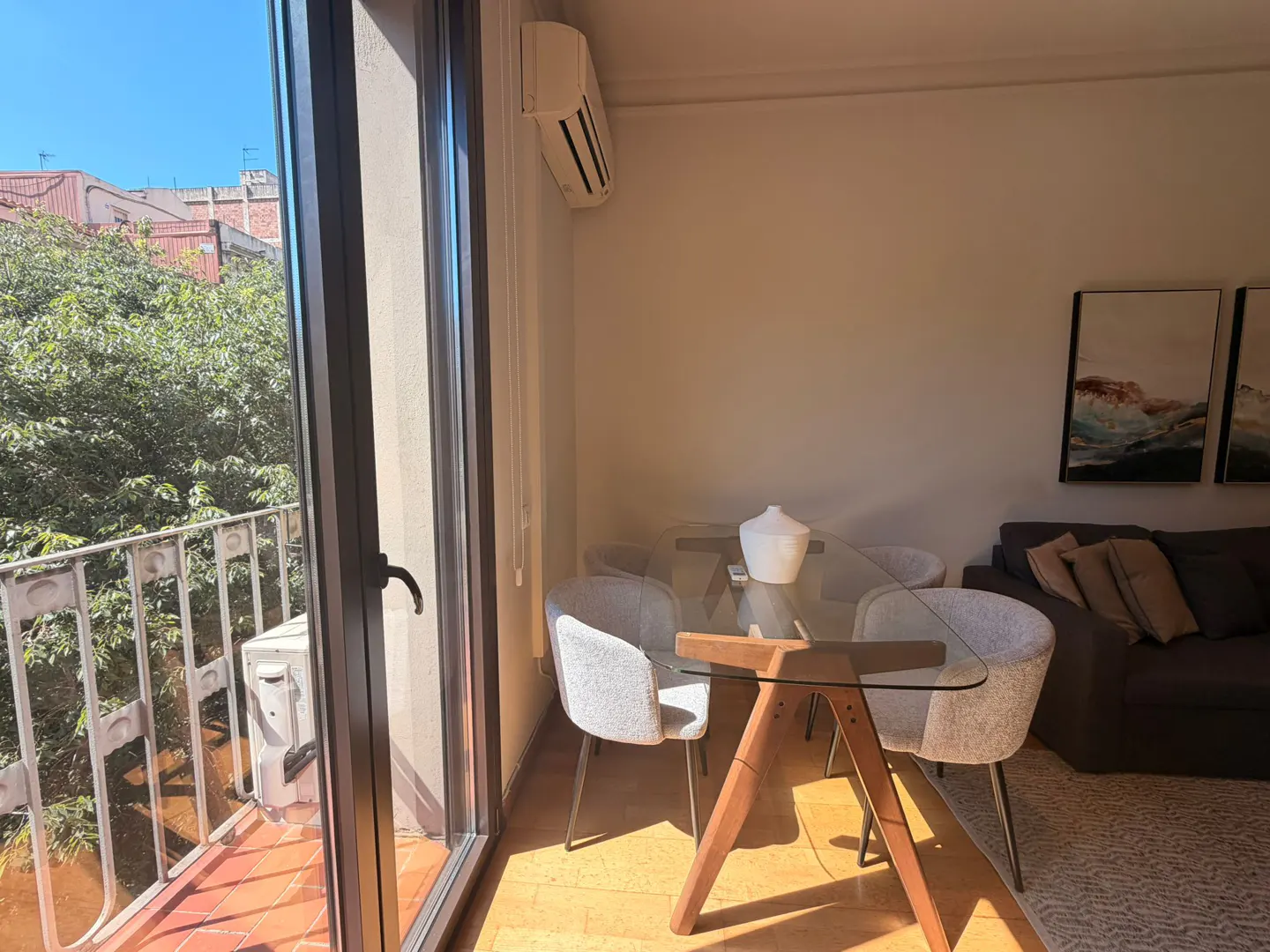 Bright living room with glass table, gray chairs, and a brown sofa. Balcony with green trees visible through the open door.