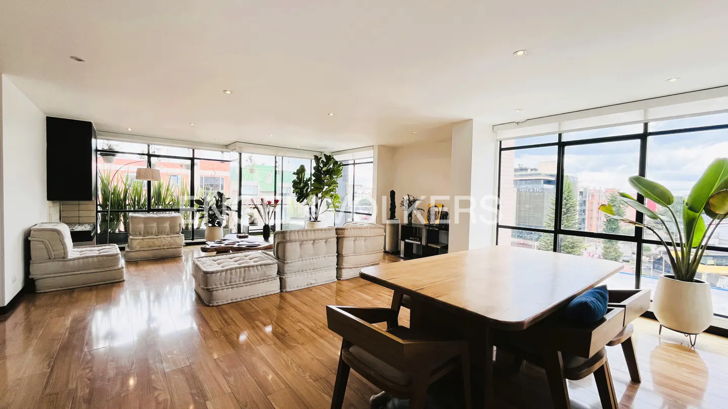 Bright, open living space with wood floors, modular seating, and large windows overlooking a city. A wooden dining table is in the foreground.