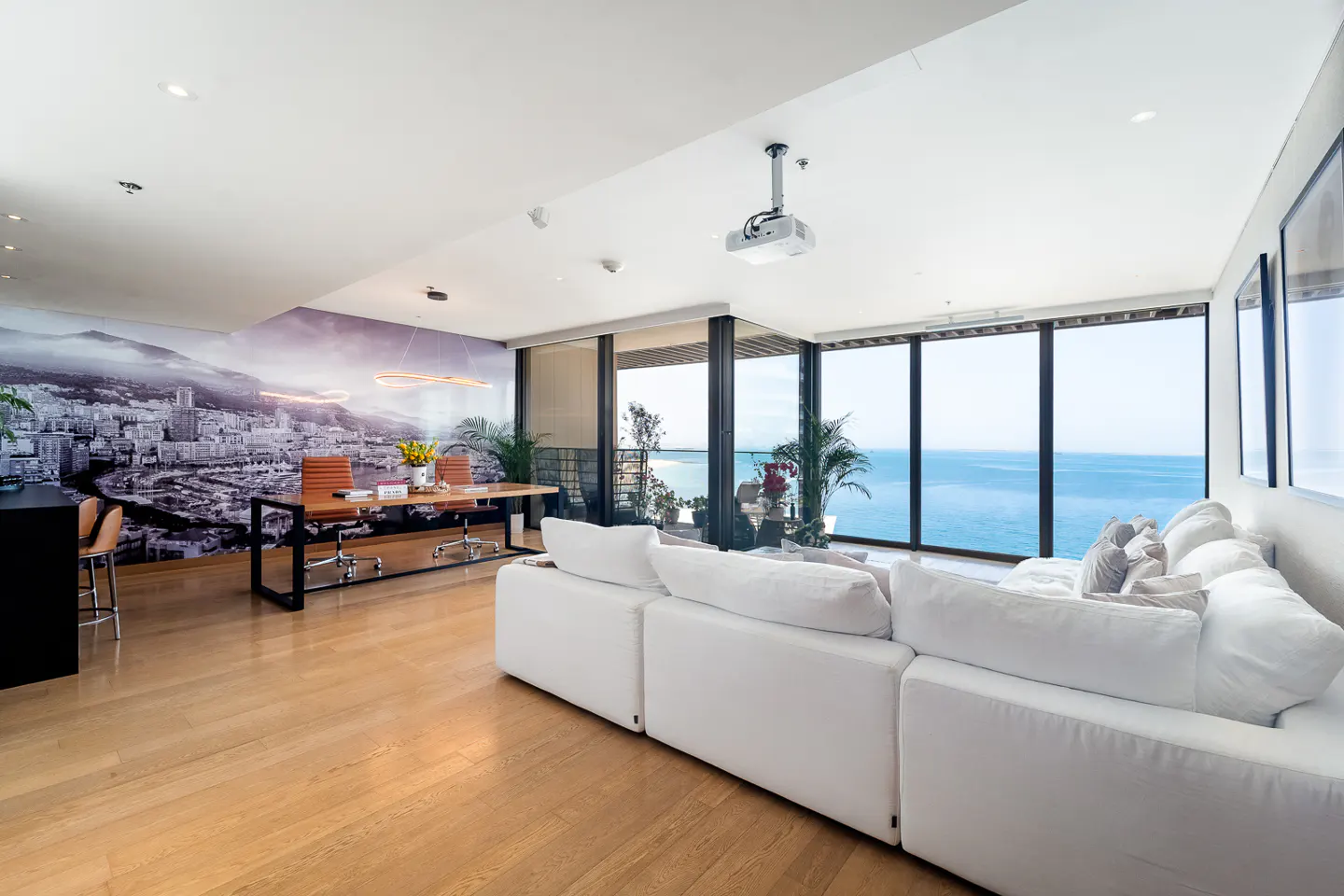 Bright, modern living room with a white sectional sofa, wood floors, and a large window overlooking the ocean. A desk and city mural are in the background.