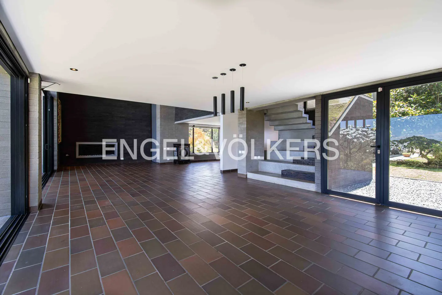 A spacious, modern living room with brick floors, a black brick wall, and large windows. Concrete stairs lead to the upper level.
