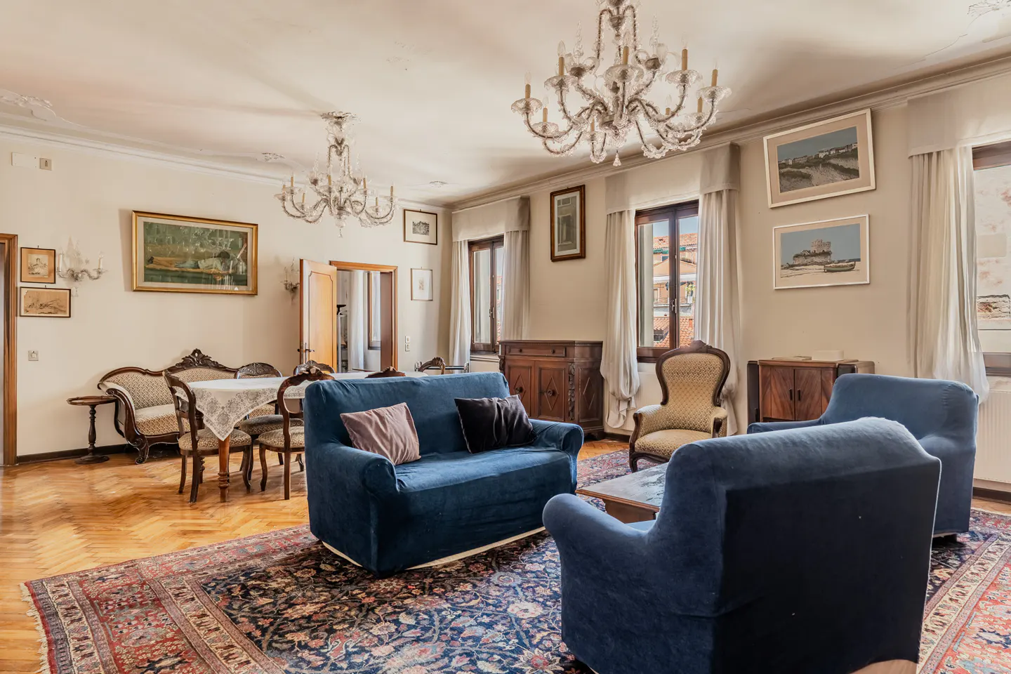 Elegant living room with blue sofas, ornate chandeliers, a patterned rug, and vintage furniture on hardwood floors.