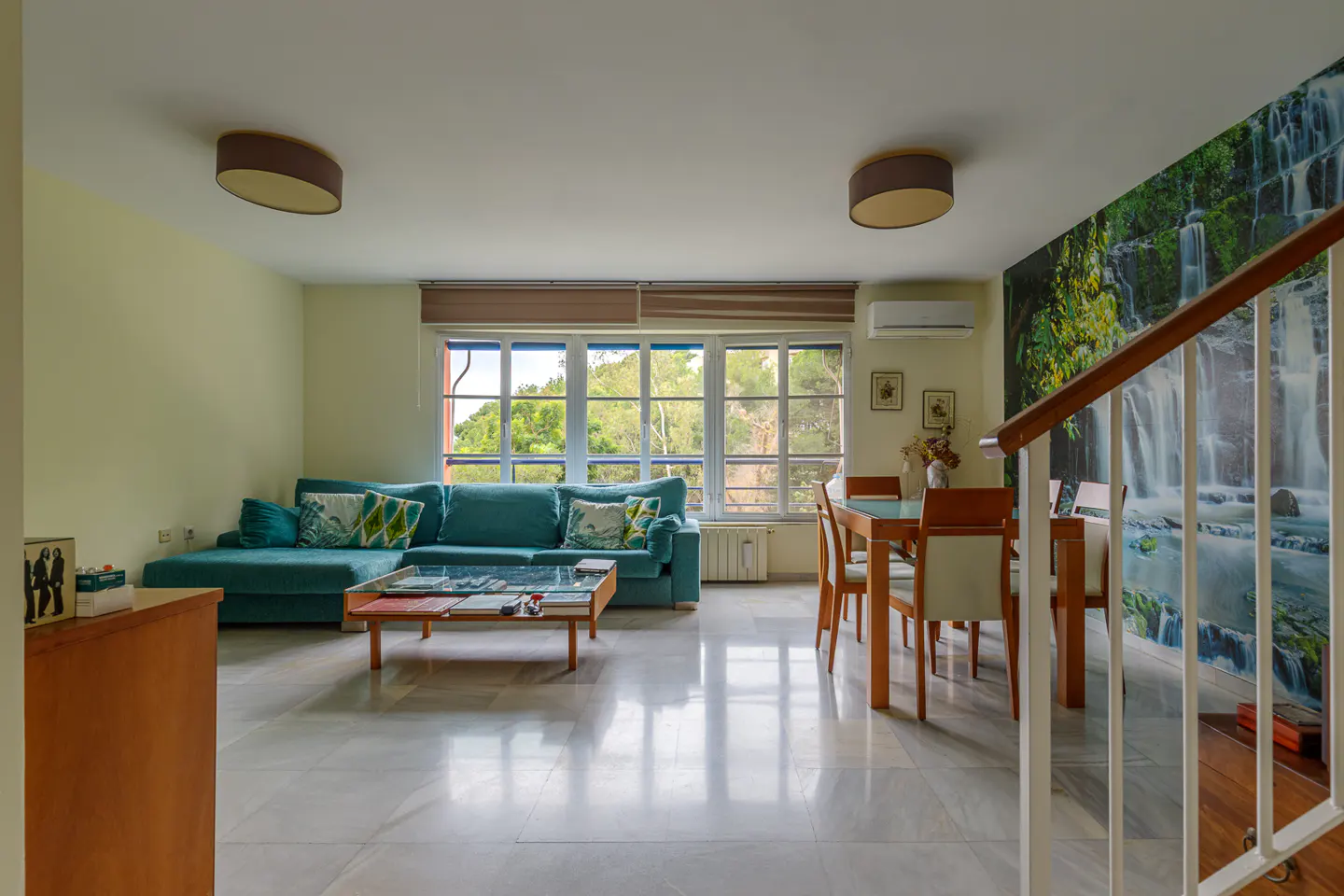 Bright living room with a teal sofa, wood table, and dining set. A large window and waterfall mural add natural elements.
