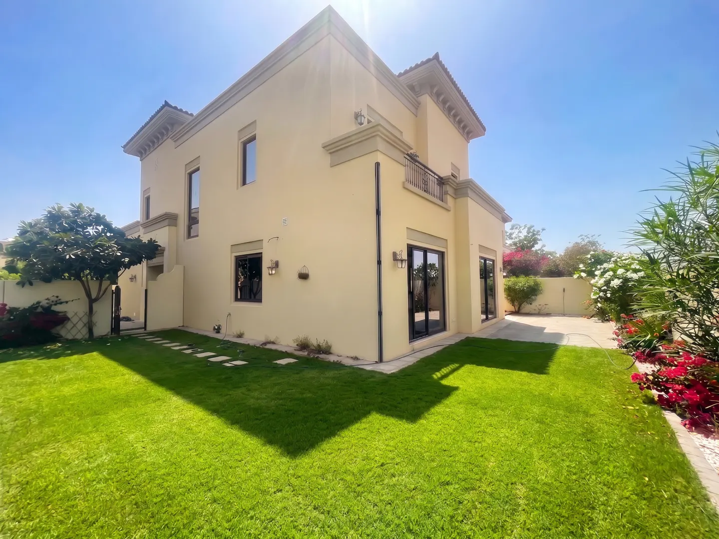 Two-story beige house with a green lawn, black framed windows, and a small balcony under a clear blue sky.