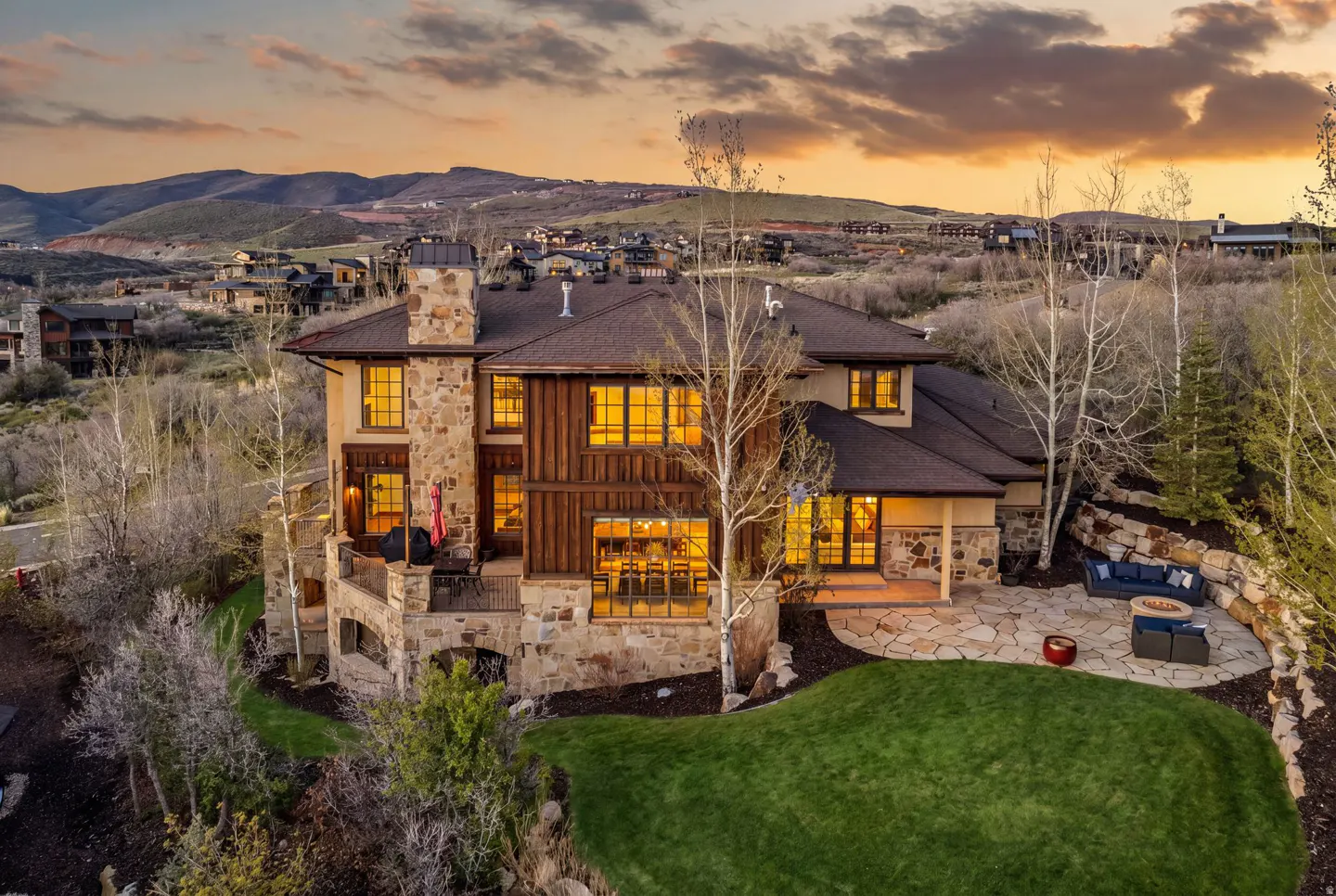 Exterior view of a luxury home with stone and wood facade, patio, and mountain views at sunset.