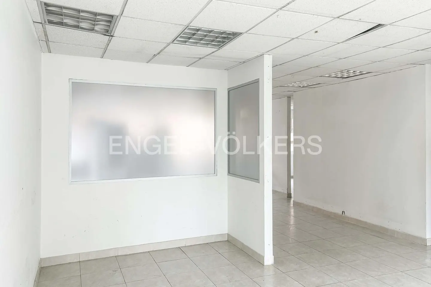 Empty office space with white walls, tile floor, and frosted glass panels. Ceiling has recessed lighting. "Engel & Völkers" logo on the glass.
