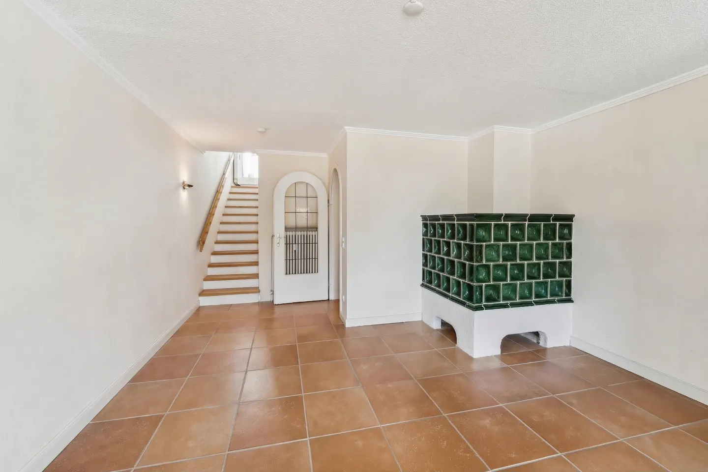 Bright room with terracotta tile floors, a green tiled masonry heater, and a staircase leading to an arched doorway.