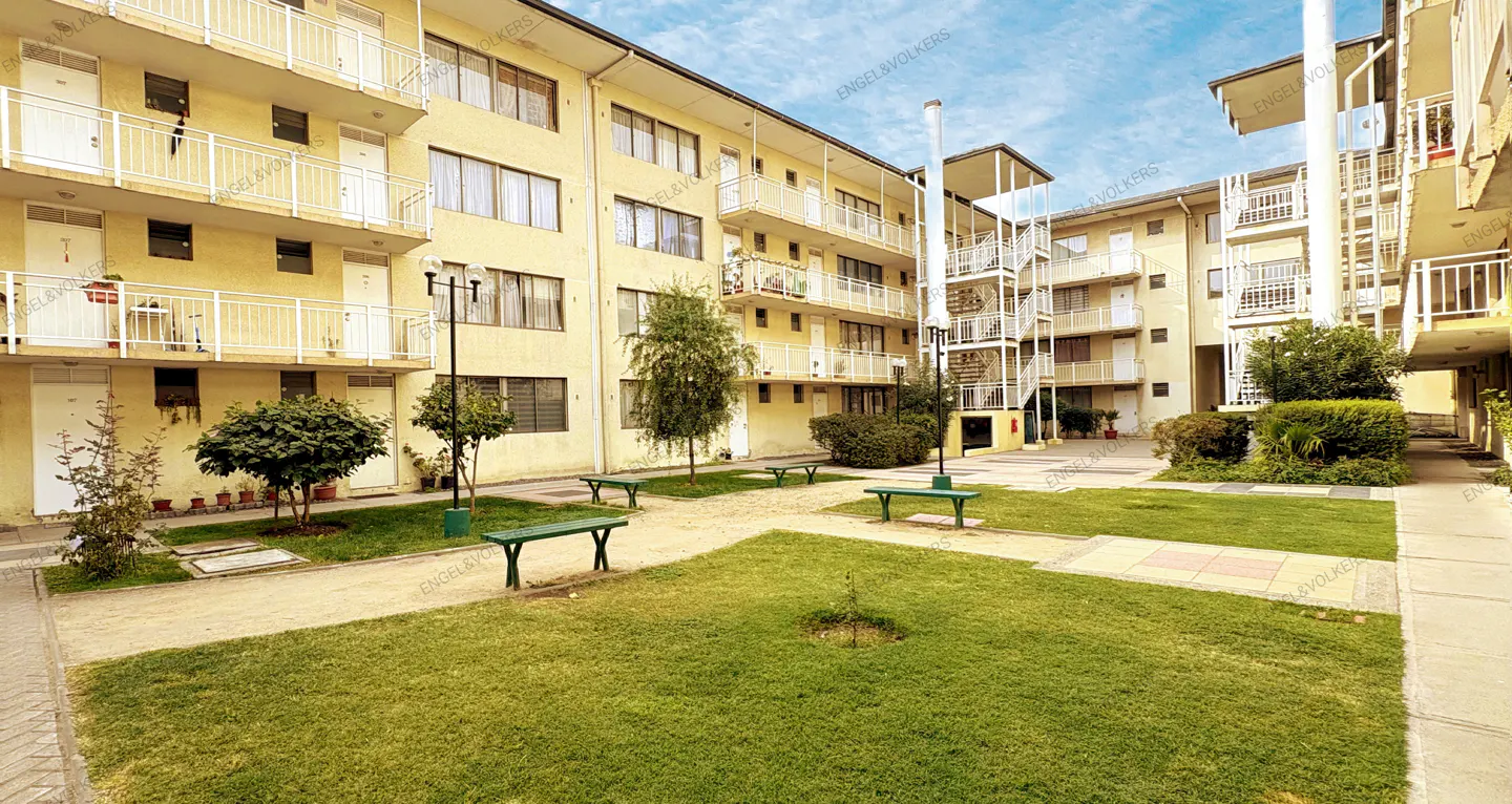 Exterior view of a yellow apartment complex with a green courtyard, benches, and trees under a blue sky.