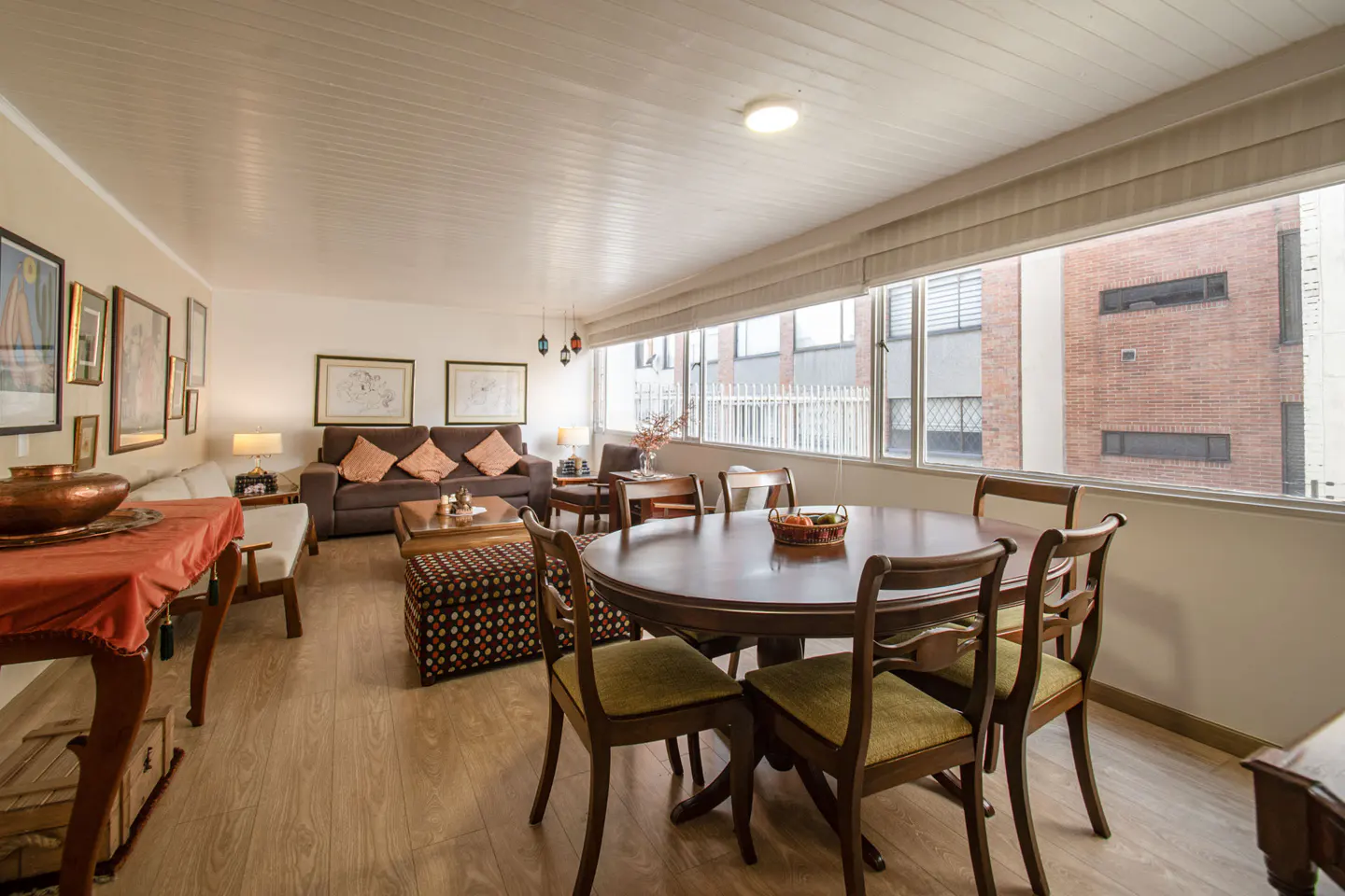 Bright living room with wood floors, a brown sofa, and a round dining table with chairs near a large window.