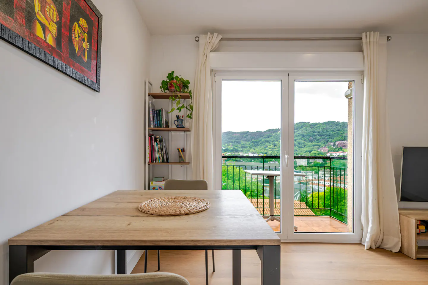 Bright dining room with a wooden table, chairs, and a view of green hills through a balcony door. A bookshelf and artwork add character.