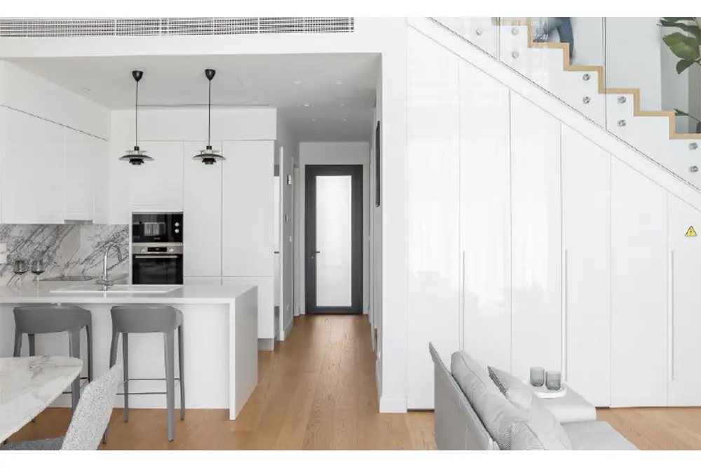 Bright, modern interior with white cabinets, marble backsplash, and wood floors. Gray stools at the island and stairs with glass railing.