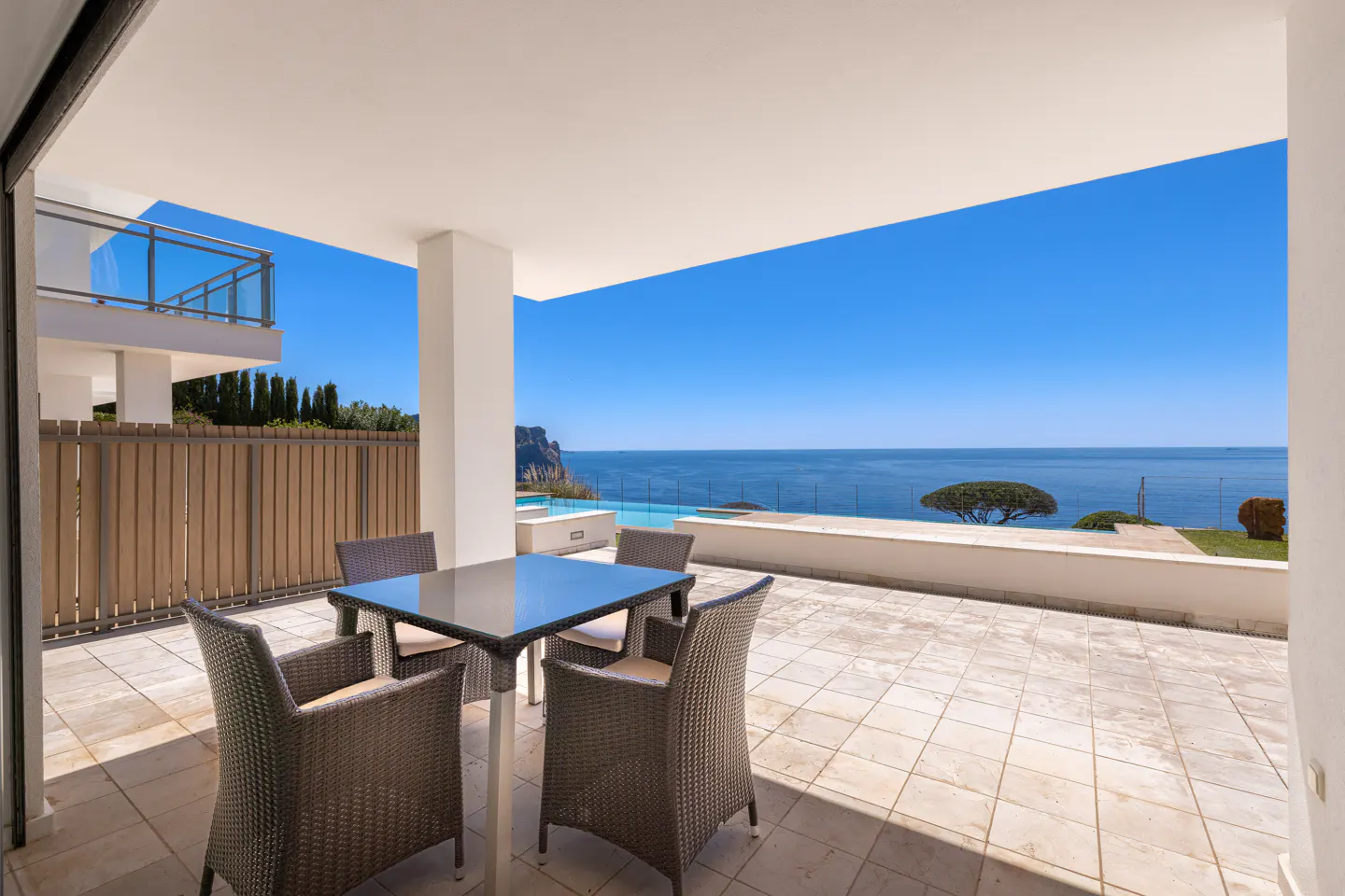 Outdoor patio with wicker chairs and glass table overlooking the ocean on a sunny day.