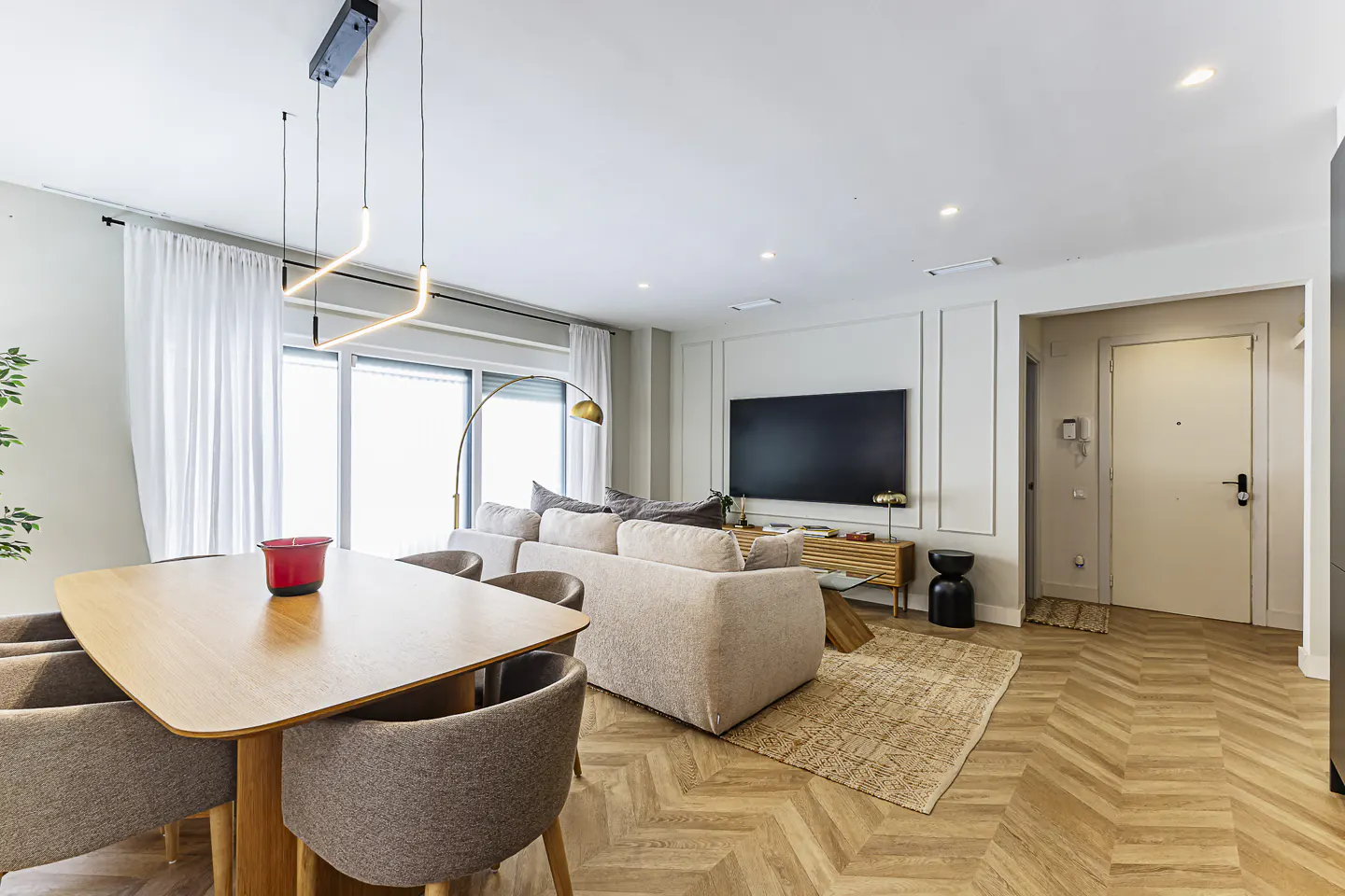 Bright living room with a dining table, beige sofa, TV, and herringbone wood floors. White walls and curtains.