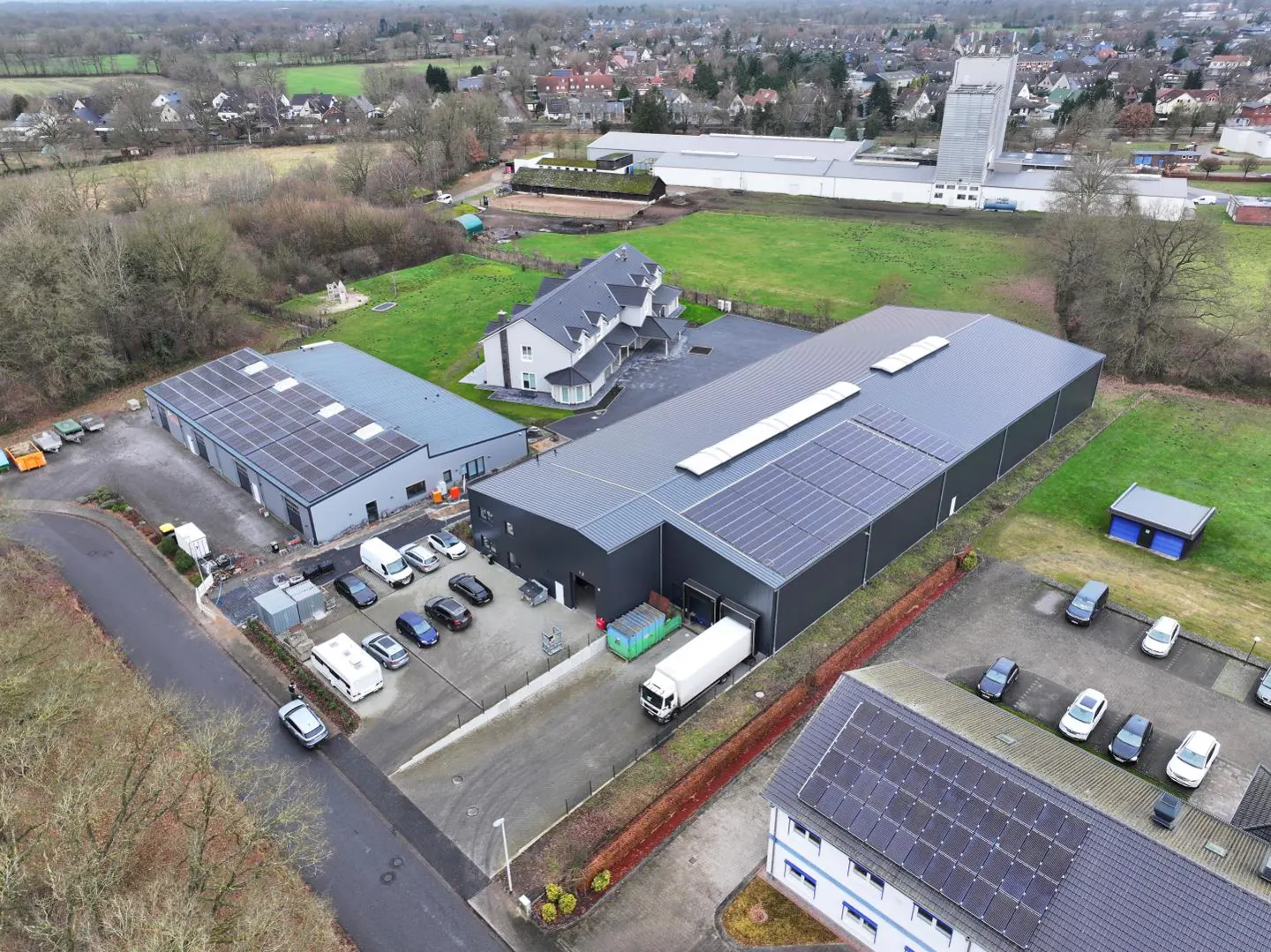 Aerial view of a modern, gray industrial building with solar panels, surrounded by greenery and parked vehicles.