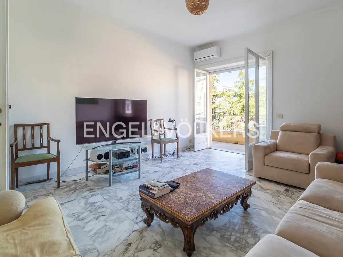 Bright living room with marble floors, beige sofas, and a dark wood coffee table. Balcony doors open to greenery. A TV displays "Engel & Völkers" logo.