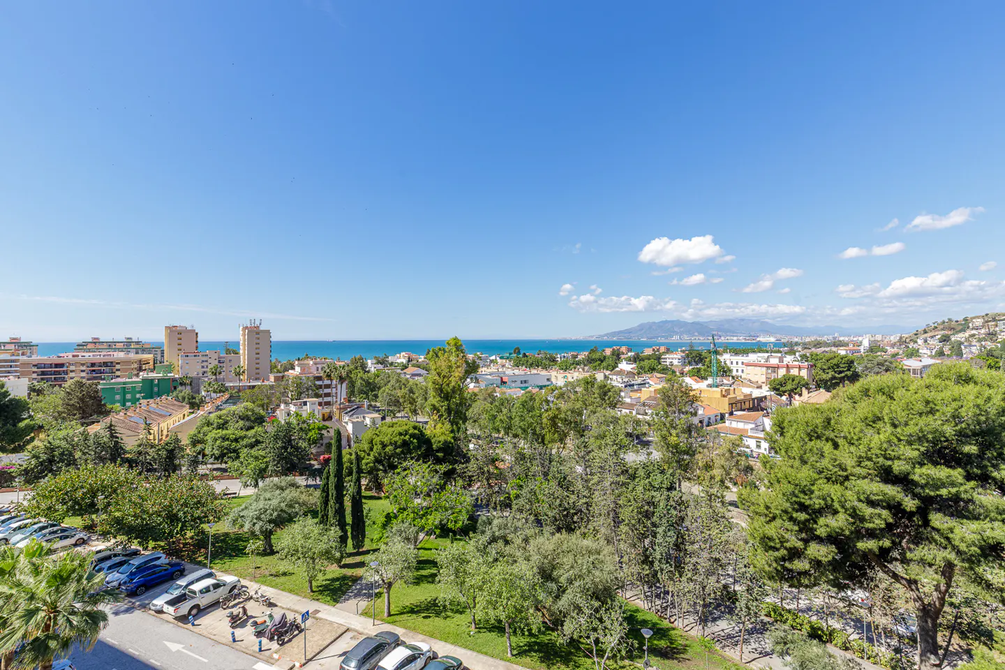 A high-angle view shows a city with buildings, trees, and a blue ocean under a clear blue sky. A parking lot with cars is in the foreground.