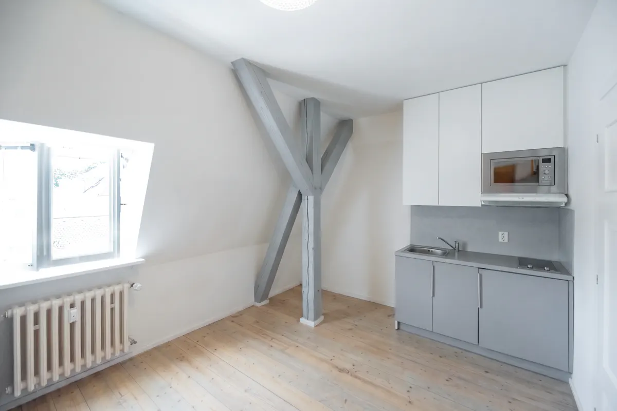 Modern attic kitchen with light wood floors, grey cabinets, white upper cupboards, and exposed grey structural beams.