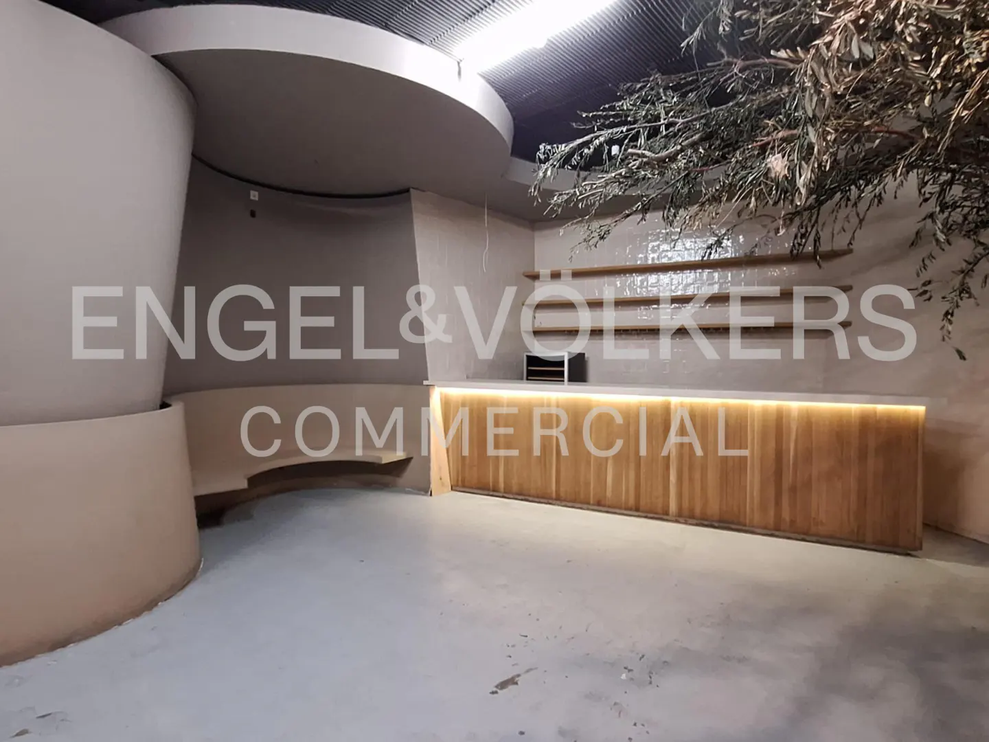 Commercial space with a light wood bar, curved seating, and decorative branches. The walls are light beige, and the floor is gray.