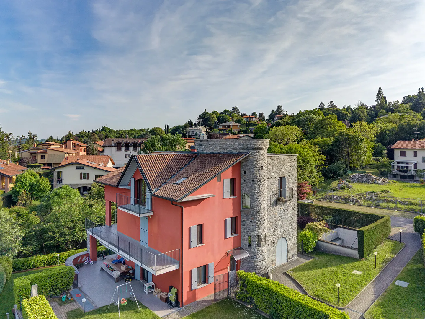Aerial view of a red villa with a stone tower, tiled roof, balconies, and a lush garden in a scenic hillside neighborhood.