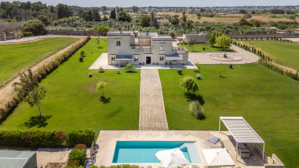 Aerial view of a light-stone villa with a pool, patio, and lush green lawn. A stone path leads to the house. Trees and fields in the background.