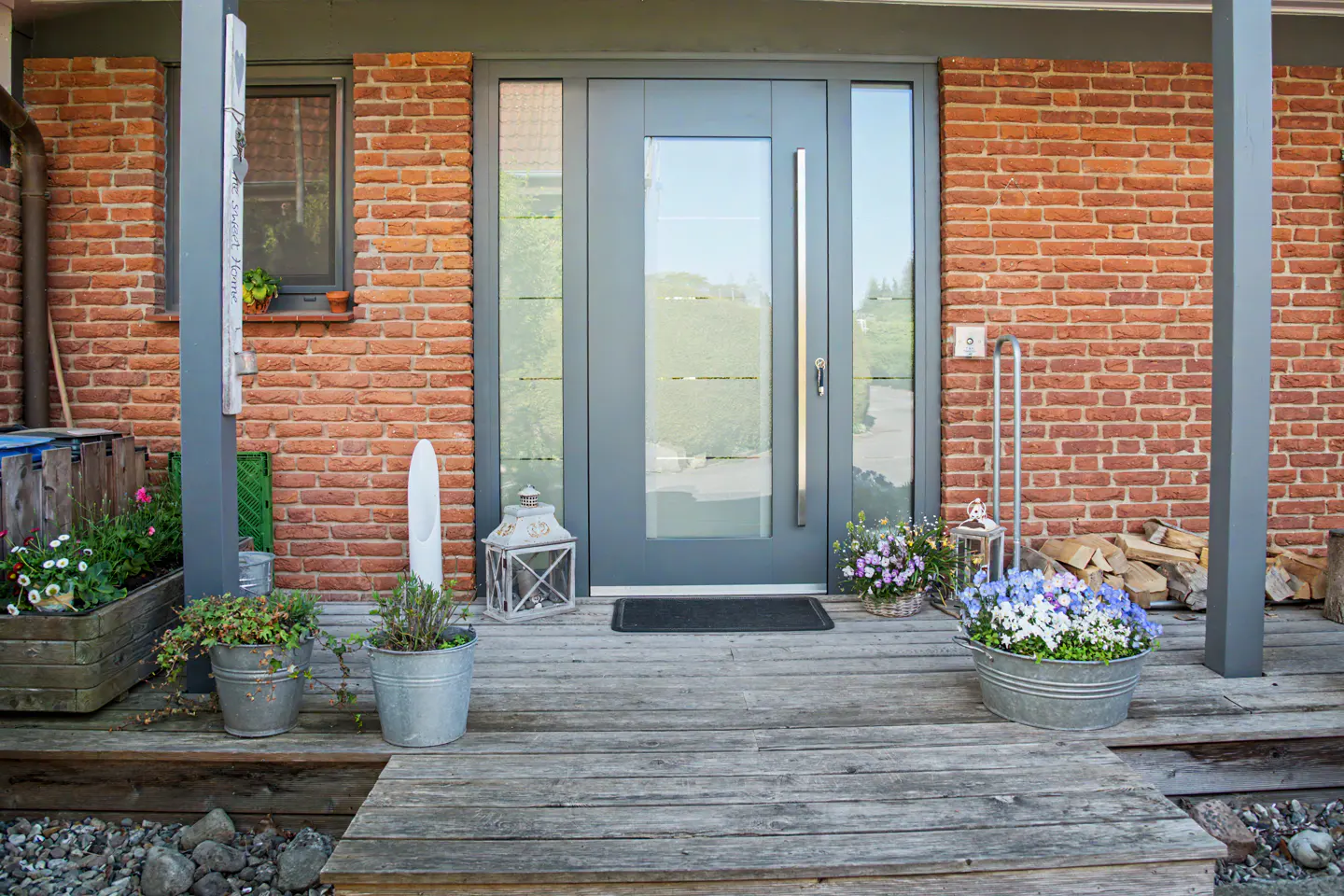 Front entrance of a brick house with a gray door, wooden porch, and potted plants.