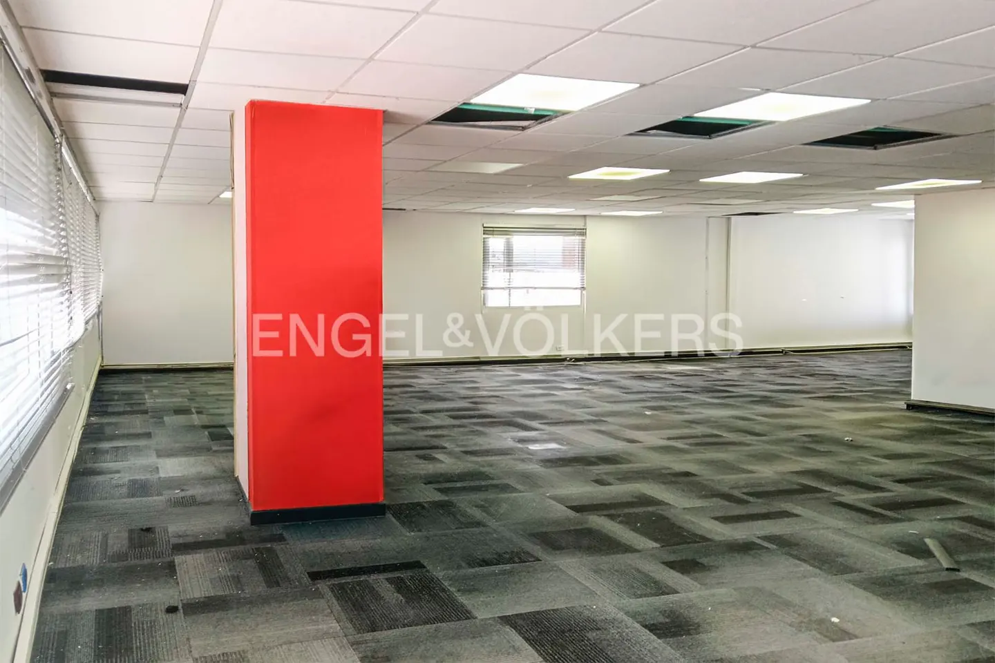 Empty office space with gray carpet, white walls, and a red support beam. Windows with blinds are visible. Engel & Volkers logo is superimposed.